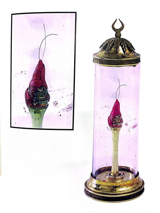 Photograph of a hair-relic reliquary in a cylindrical glass display case.